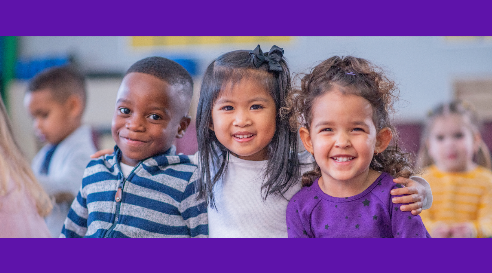 Three small children are sitting next to each other in a classroom. They are smiling. The girl in the middle has her arms around the shoulders of the other two.