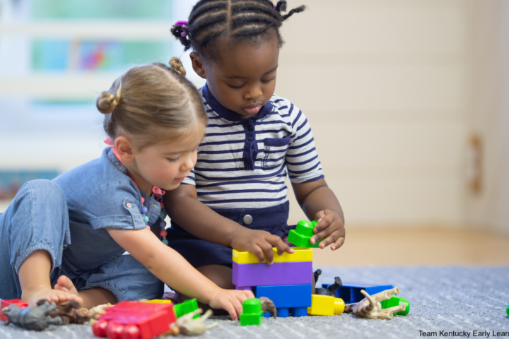 Two toddlers playing with plastic building blocks on a carpet.